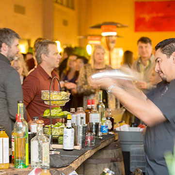 Photograph from behind a bar while patrons watch a bartender make drinks